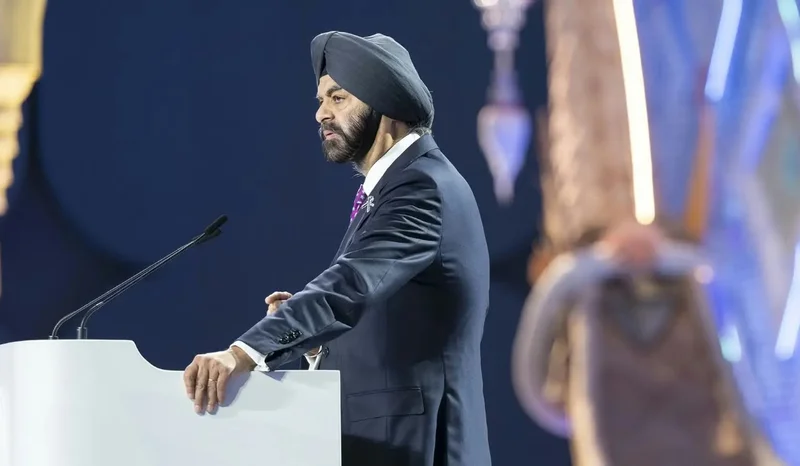 Ajay Banga, the President of the World Bank Group, is captured delivering a speech from behind a white podium. He is dressed in a formal suit and a dark turban, set against a deep blue background at an international event.