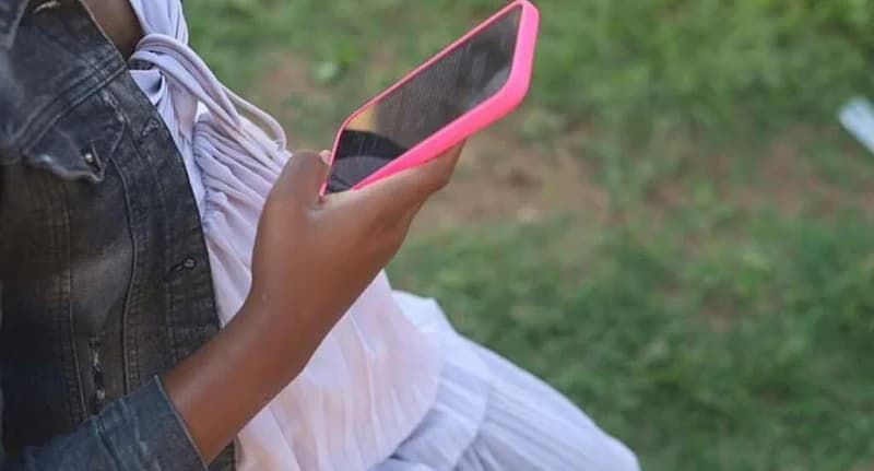 A close-up photograph shows a person holding a smartphone with a distinctive pink protective case. The individual, partially visible, wears a denim jacket over a white top, set against a blurred green outdoor background.