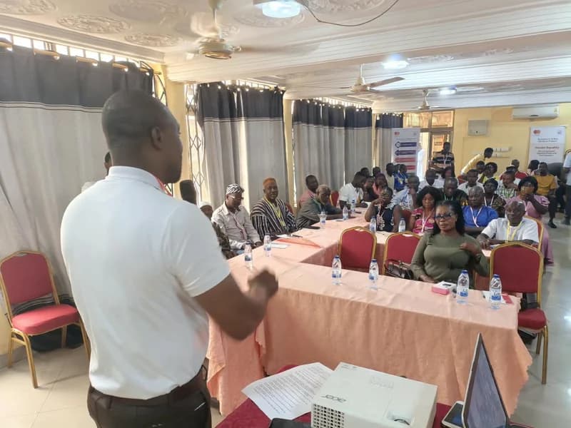 A man in a white polo shirt addresses a diverse group of participants during a business management training workshop. The audience is seated at U-shaped tables covered in pink cloths, with water bottles and training materials present in what appears to be a conference hall in Techiman.