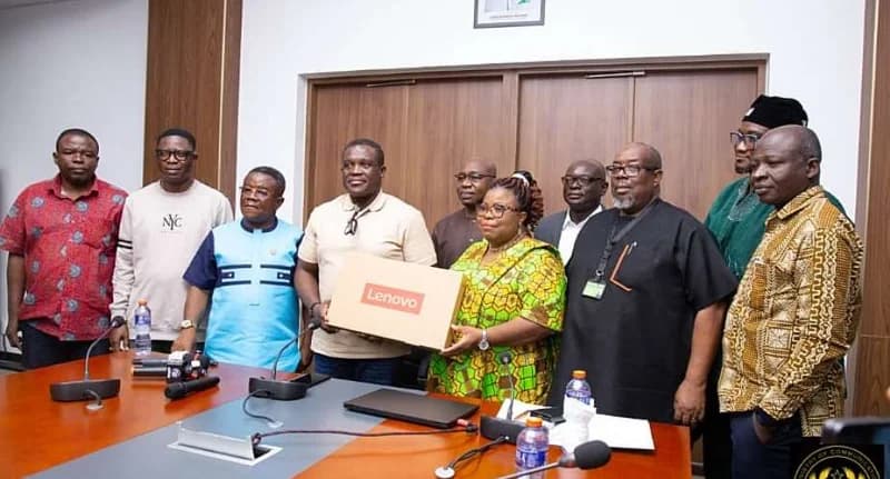 A group of officials, including Ghana's Minister for Communications and Digitalisation, Ursula Owusu-Ekuful, are pictured during a formal handover ceremony. Two individuals in the center are holding a Lenovo laptop box over a conference table equipped with microphones, signifying the distribution of digital tools for education.