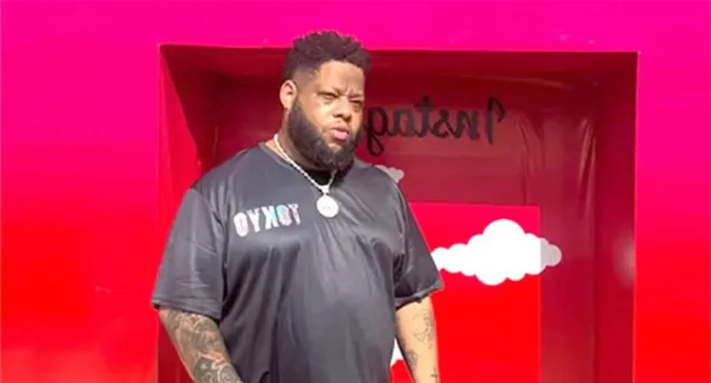 Ghanaian musician and entrepreneur D-Black is pictured standing in front of a vibrant pink background. He is wearing a black t-shirt with the word 'TOKYO' printed on it and a prominent silver chain with a medallion.