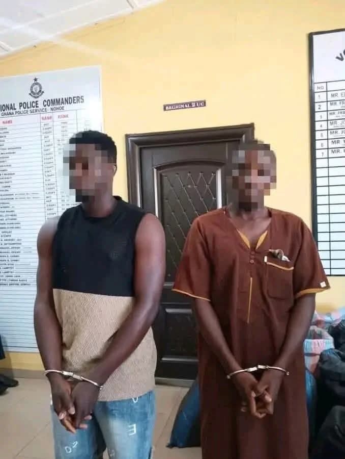 Two male suspects are seen standing in handcuffs inside a Ghana Police Service office. Their faces are blurred, and they are positioned in front of a door and information boards listing police commanders. The man on the left wears a two-tone sleeveless shirt and jeans, while the man on the right is in a brown traditional kaftan.