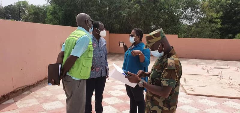 A group of four officials, including a Ghanaian military officer and an individual in a high-visibility safety vest, are gathered at an outdoor site for an inspection. They are wearing face masks and appear to be discussing technical details near concrete slabs that likely serve as access points for an underground biodigester system.