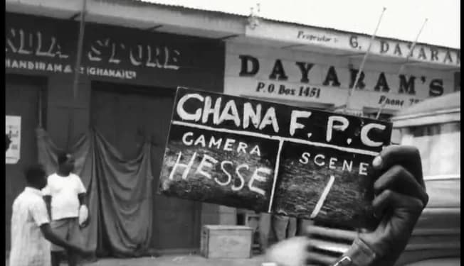 This black and white archival photograph captures a moment from a historical film production in Ghana, showing a hand holding a clapperboard labeled 'GHANA F. P. C.' in the foreground. The background features vintage shopfronts such as 'Dayaram's' and 'Handiram & (Ghana) Ltd' with two men standing near a doorway.