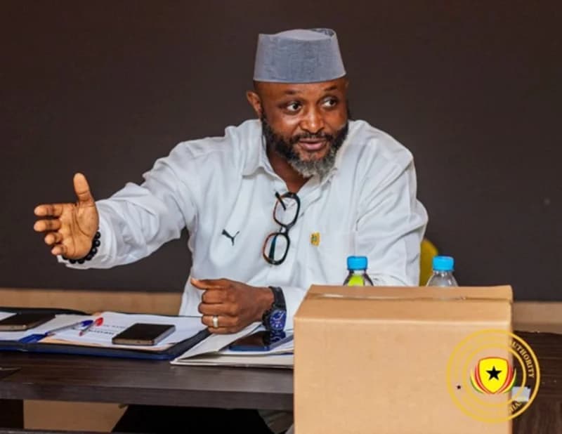 The image shows Mustapha Ussif, Ghana's Minister for Youth and Sports, seated at a table during what appears to be a formal meeting or press briefing. He is gesturing with his right hand while looking towards someone off-camera, with documents, mobile phones, and water bottles arranged on the table in front of him.