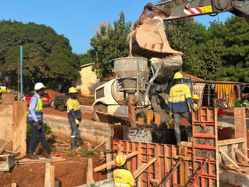 Construction workers are seen actively working on a road infrastructure project, involving concrete pouring and the use of heavy machinery like an excavator and a concrete mixer. The workers are wearing high-visibility gear and hard hats as they manage formwork on a site surrounded by lush greenery and local buildings.