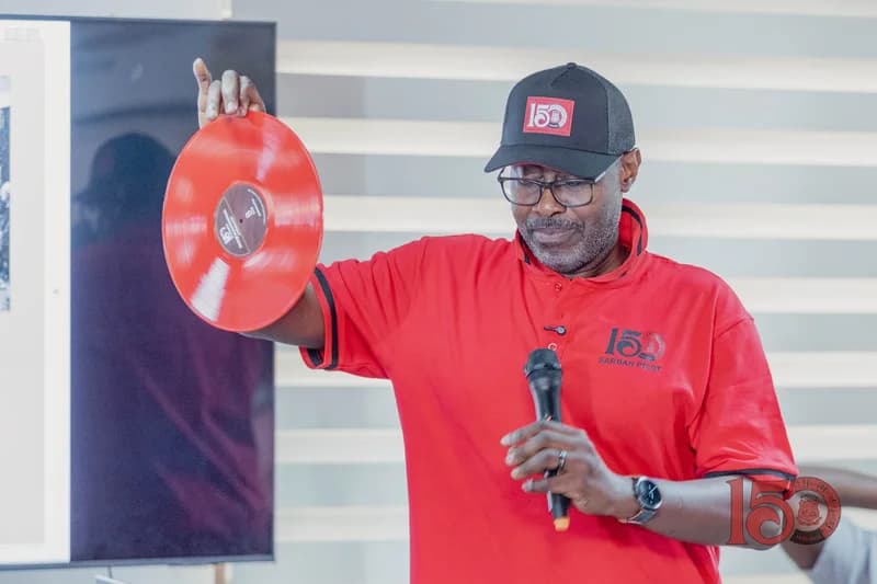 A man wearing a red polo shirt and a black baseball cap with commemorative branding holds up a red vinyl record and a microphone. He appears to be speaking at an event related to the 150th anniversary of Mfantsipim School, as indicated by the 'Sarbah Picot' house name and '150' logo on his clothing.