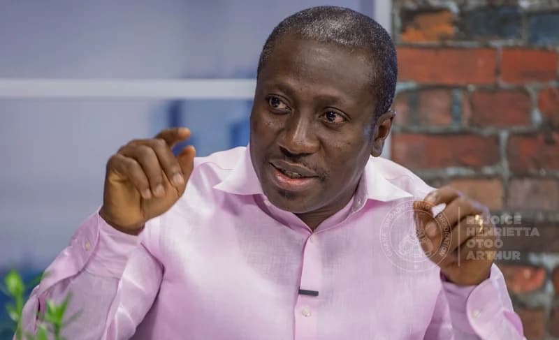 A close-up photograph of Ghanaian politician Alexander Afenyo-Markin wearing a light pink shirt while gesturing with his hands during a media appearance. He is set against a background featuring a brick wall and studio lighting, with a visible watermark from the Ghana Broadcasting Corporation.