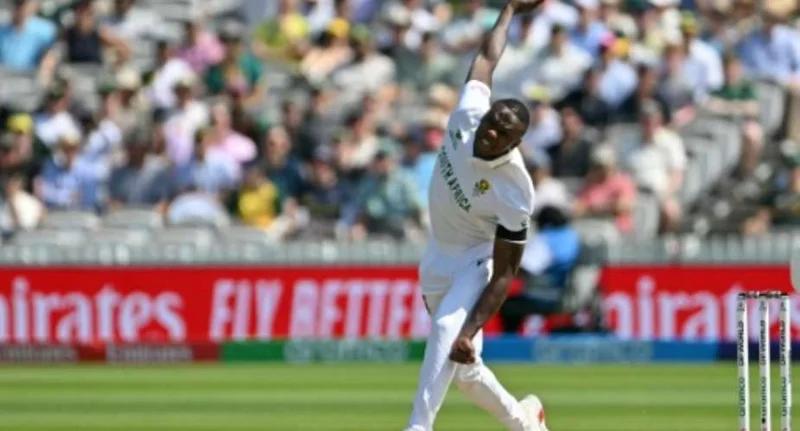 South African fast bowler Kagiso Rabada is captured in action during a cricket match, delivering a ball in his white Test kit. The background features a crowded stadium and advertising hoardings for Emirates and Aramco.