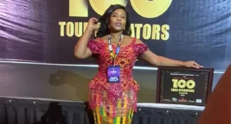 Mary Asamoah poses with her Africa Travel 100 award at the Akwaaba travel market event. She is dressed in a vibrant red and yellow kente-style outfit, standing in front of a black backdrop featuring event branding.