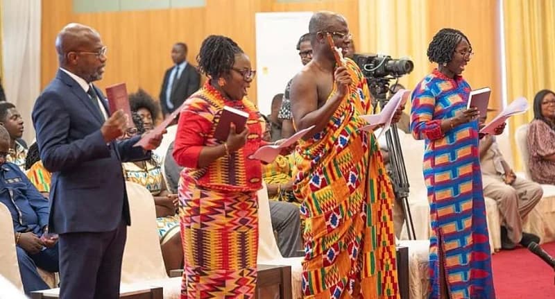 Four newly appointed officials are seen standing in a row while taking an official oath of office. They are holding religious texts and documents, dressed in a mix of formal Western suits and colorful traditional Ghanaian Kente cloths during a state ceremony.