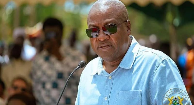 President John Dramani Mahama is captured speaking at a public event, wearing a light blue button-down shirt and dark sunglasses. A microphone is positioned in front of him, and the seal of the Presidency of Ghana is visible on his left sleeve.