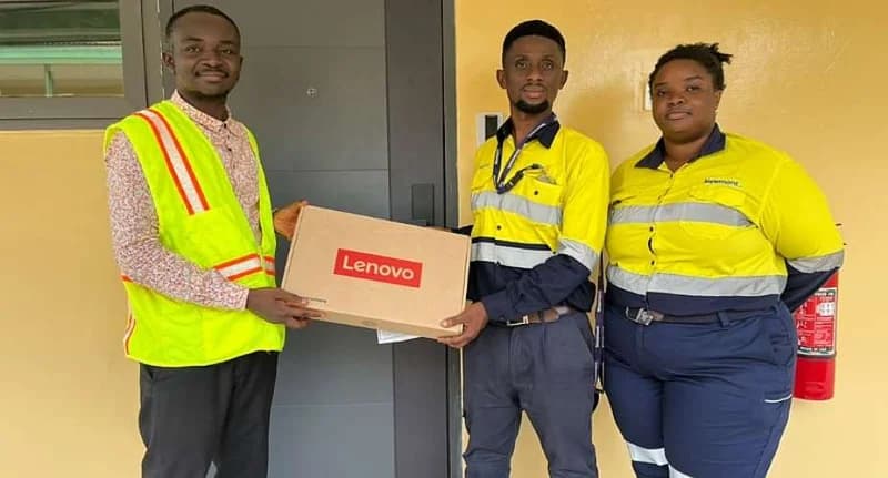 Three individuals are pictured during a presentation ceremony. Two Newmont employees in high-visibility uniforms are handing a Lenovo laptop box to another man wearing a reflective vest. This event signifies a corporate donation or award presentation.