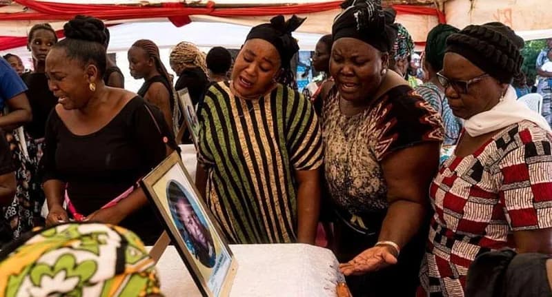 A group of women are captured in a moment of intense grief, mourning around a framed photograph of a deceased individual. The scene appears to be a memorial or funeral service held under a red and white canopy, with several women wearing traditional headwraps and expressing deep emotional distress.