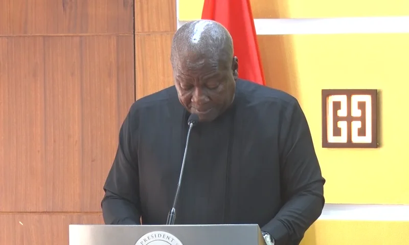 President John Mahama is captured delivering a formal address from behind a silver podium. He is dressed in a black long-sleeved outfit, standing against a backdrop featuring a yellow wall with a Gye Nyame symbol and a red flag.
