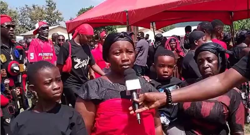 A woman dressed in traditional red and black Ghanaian mourning attire speaks into a microphone during a public gathering. She is surrounded by somber-looking community members, including young children, under a large red canopy, indicating a funeral or a protest for justice.