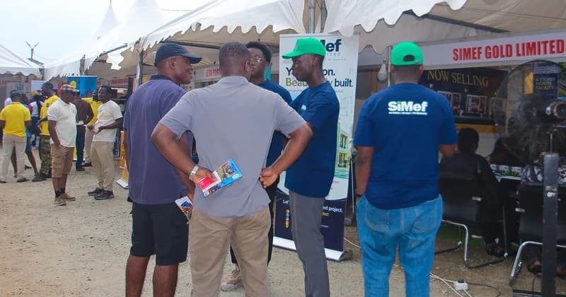 Patrons and exhibitors interact at a housing exhibition booth during the Republic Bank–JoyNews Habitat Fair in Ho. Several individuals are seen wearing branded SiMef apparel while discussing real estate options under outdoor event tents.