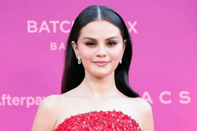 A portrait of American singer and actress Selena Gomez posing against a vibrant pink background. She is wearing a red sequined top and silver hoop earrings, with her dark hair styled straight and tucked behind her ears. The background features logos including Afterpay and BATCH.