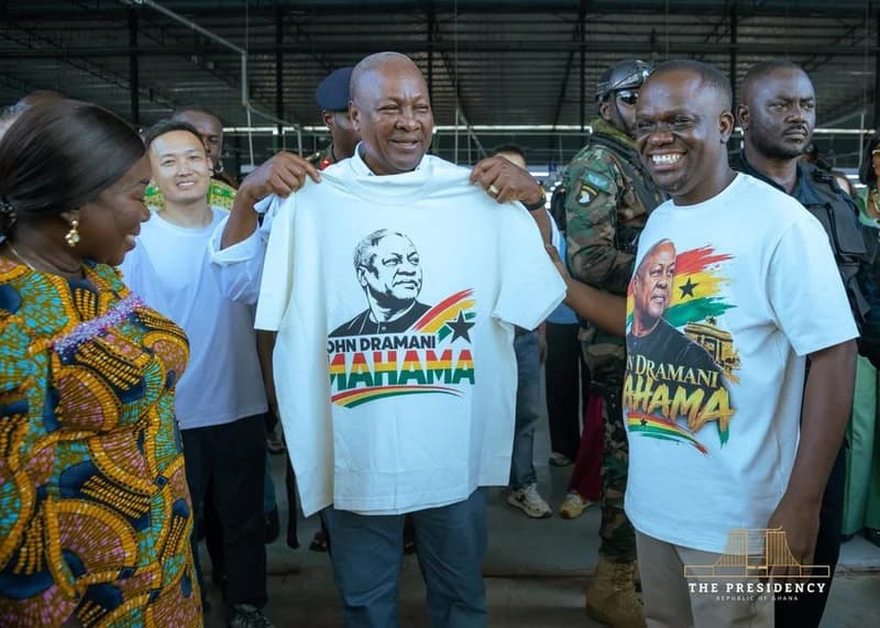 President John Dramani Mahama is seen holding up a white t-shirt featuring his portrait and name during a visit to an industrial facility. He is accompanied by security personnel and individuals wearing similar branded apparel within what appears to be a factory setting.