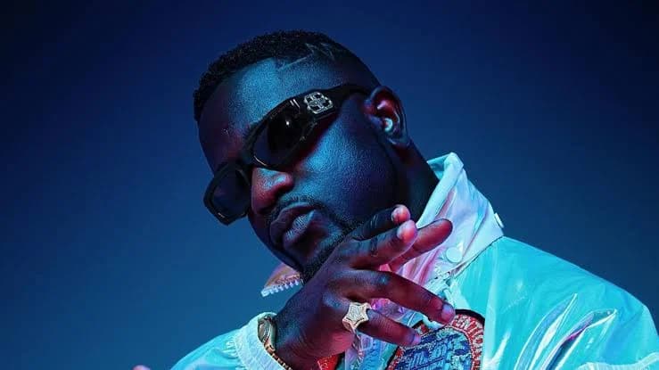 A close-up photograph of prominent Ghanaian rapper Sarkodie posing in stylish sunglasses and a white reflective jacket. He is seen wearing a large gold ring and a watch, set against a dark blue background with dramatic lighting.