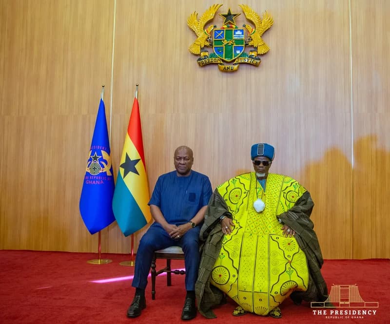 President John Mahama is seated next to a high-ranking traditional ruler, likely the Regent of Dagbon, during a formal state visit. They are positioned in front of the Ghana coat of arms and national flags, symbolizing a meeting between government and traditional authority.