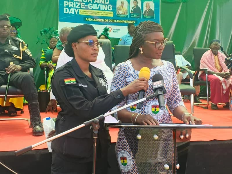 A woman in an Assisi Girls' Senior High School commemorative cloth speaks at a podium during a Speech and Prize-Giving Day event. She is flanked by a female Ghana Police officer in a black uniform, with a Joy News microphone visible as she addresses the audience. The background features a banner celebrating 67 years of the school and the launch of its 70th-anniversary celebrations.