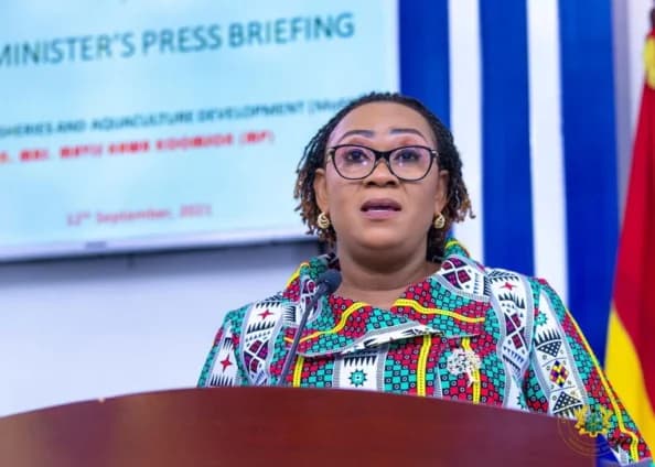 A photograph of Ghanaian politician Mavis Hawa Koomson speaking at a podium during a press briefing. She is wearing a vibrant, multicolored patterned dress and eyeglasses, with a digital screen and the flag of Ghana visible in the background.