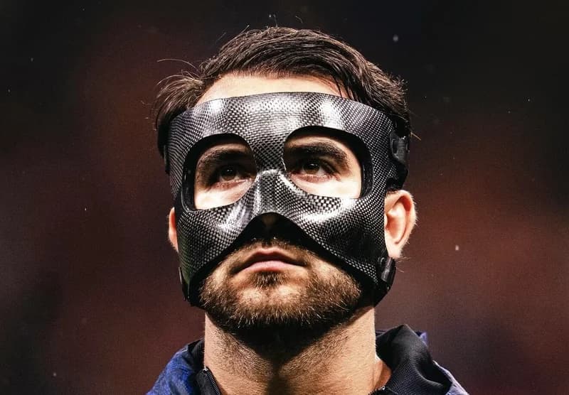A close-up photograph of a professional football player wearing a black carbon-fiber protective facial mask. The subject is looking upwards with a serious expression, highlighting the specialized equipment often used by athletes to play through facial injuries.