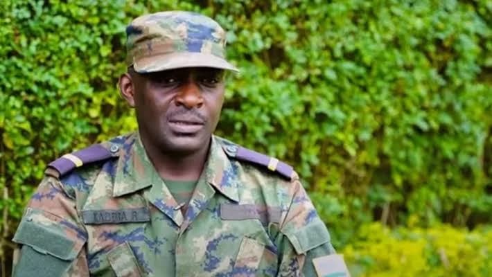 A close-up photograph of a man in a military camouflage uniform and matching cap, looking directly at the camera. He has a name tag that reads "KABERA R" on his chest, and the background consists of dense green foliage.