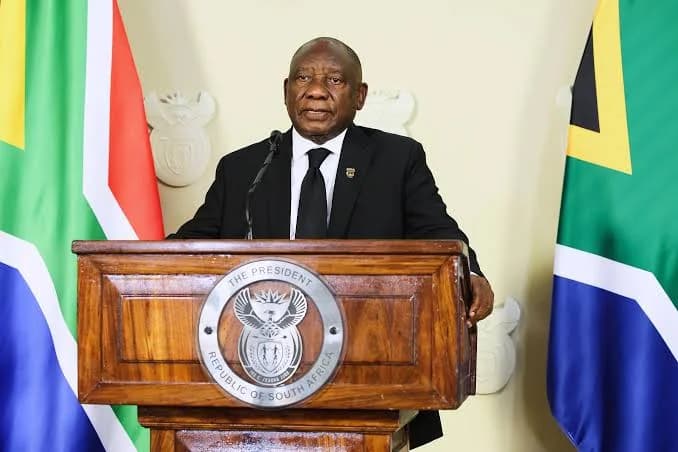 South African President Cyril Ramaphosa is seen delivering an official address from behind a wooden podium featuring the national presidential seal. He is flanked by two South African flags and stands before the national coat of arms, dressed in a formal black suit and tie. The image captures a moment of state-level communication regarding national policy and international relations.