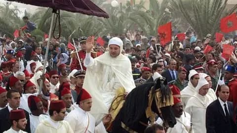 AFP via Getty Images Morocco's King Mohammed VI, wearing a long white traditional robe, rides a horse amid a large crowd of security personnel, military officers, officials and civilians, waving to the crowd.