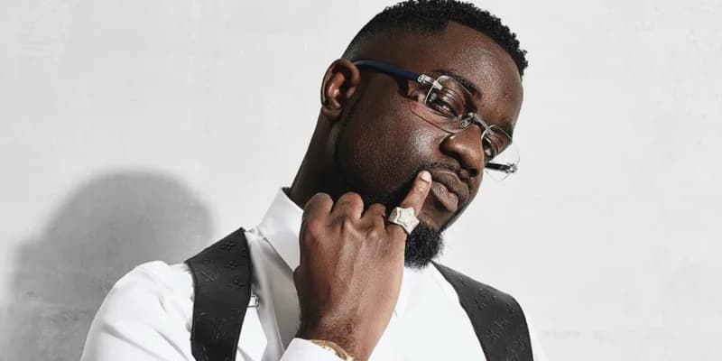 A close-up portrait of the award-winning Ghanaian rapper Sarkodie, wearing rimless glasses and a white shirt with a black monogrammed harness. He is shown in a thoughtful pose with his finger to his lips, highlighting a prominent star-shaped ring on his hand. The image is a high-quality studio photograph used to identify the artist mentioned in the news report.