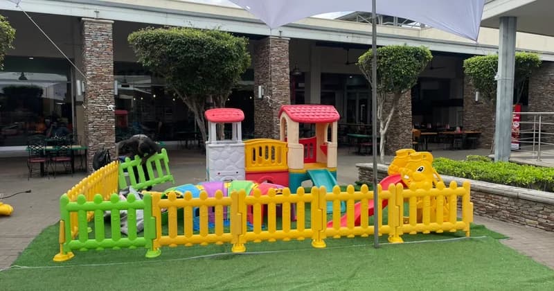 A vibrant children's play area featuring colorful plastic equipment like slides, a tunnel, and a playhouse is set up at the Accra Mall. A person is seen working on the installation, which is enclosed by a yellow and green plastic fence on artificial turf.