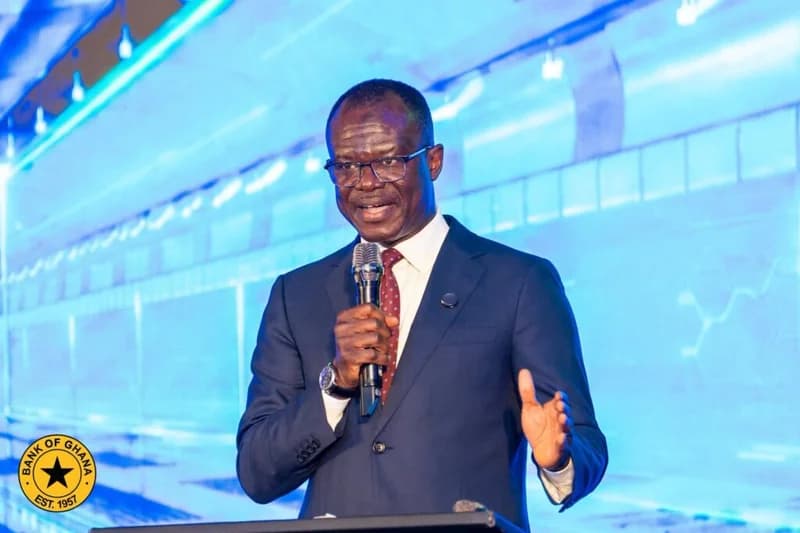 Dr. Ernest Addison, Governor of the Bank of Ghana, is seen speaking at a formal event while wearing a dark navy suit and maroon tie. He is holding a microphone and gesturing with his hand against a vibrant blue digital background featuring the Bank of Ghana logo in the corner.
