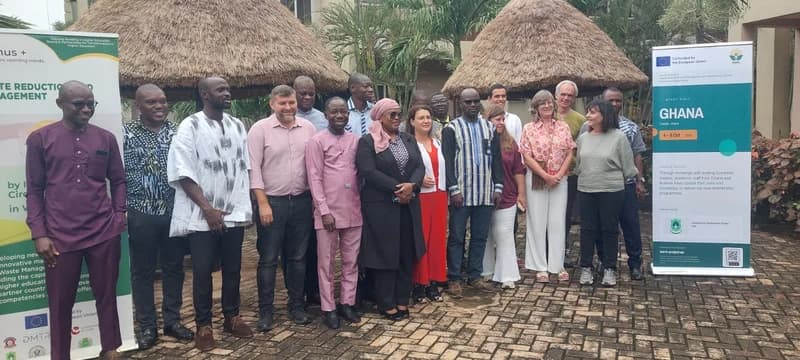 A group of academic experts and project participants pose for a group photo in Tamale, Ghana, during a study visit for the WARM project. The group is positioned between two informational banners highlighting the development of new postgraduate programmes in waste management, co-funded by the European Union and involving the University for Development Studies.