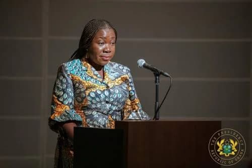 A woman in a vibrant patterned African print dress is captured speaking at a wooden podium with a microphone. The image features the official watermark of the Presidency of the Republic of Ghana in the bottom right corner, indicating a formal state function or announcement.