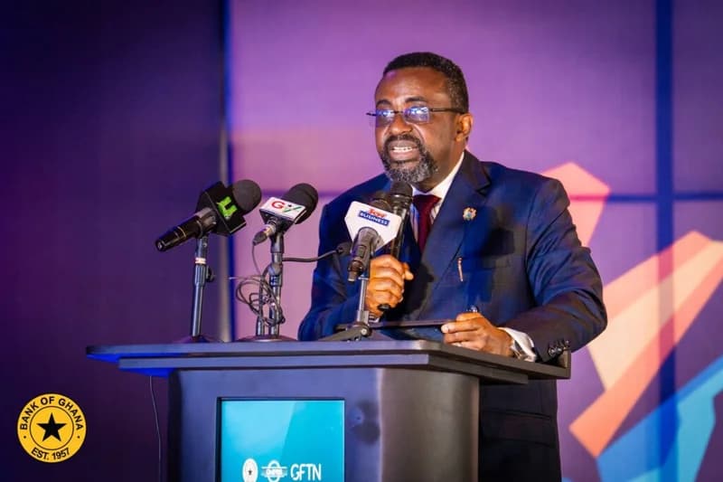 A man in a dark suit and glasses, identified as an official of the Bank of Ghana, is seen speaking at a formal event. He stands behind a podium branded with the Bank of Ghana seal and GFTN logo, surrounded by microphones from media houses like Joy Business and GTV.