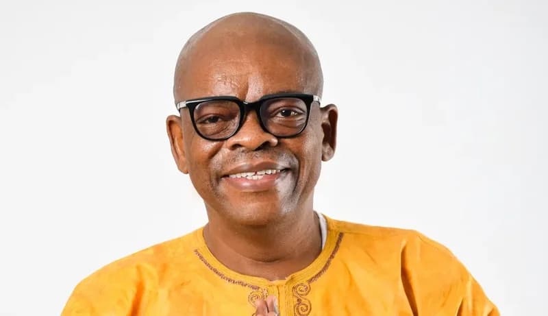 A studio portrait of Ghanaian actor and comedian Kwame Dzokoto. He is smiling warmly, wearing black-rimmed spectacles and a yellow traditional Ghanaian garment against a plain white background.