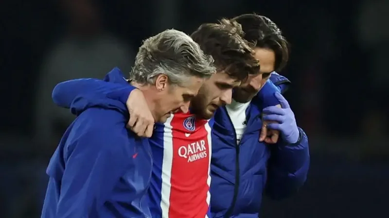 A football player wearing a Paris Saint-Germain jersey is being helped off the pitch by two medical staff members. The player appears to be in distress, aligning with reports of a significant knee injury sustained during a match.