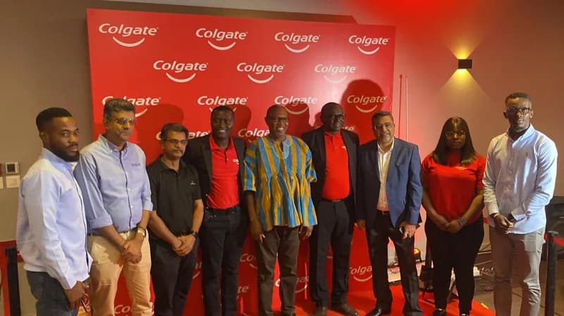 A group of nine officials and corporate representatives pose in front of a red backdrop featuring the Colgate logo. The central figure is wearing a traditional Ghanaian blue and yellow striped smock, likely representing the Ministry of Education at the scholarship campaign launch.