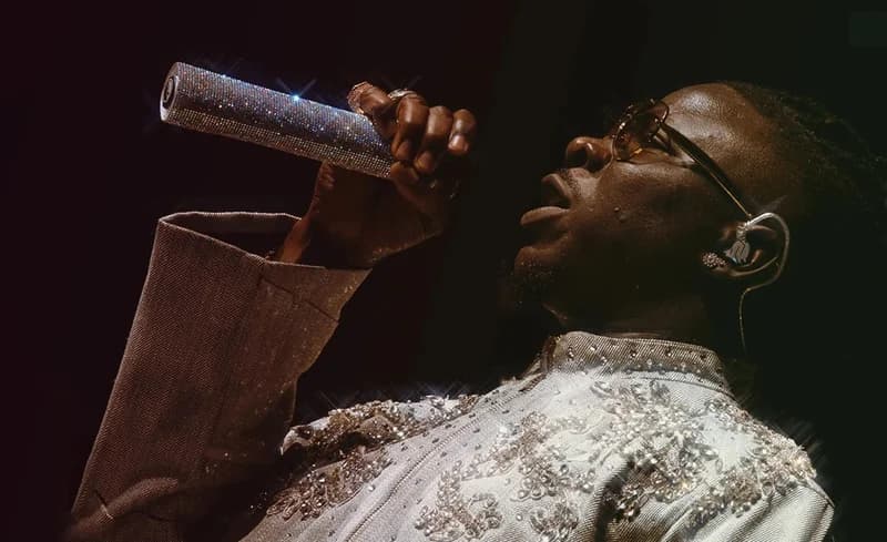 Ghanaian dancehall artist Stonebwoy is captured in a close-up profile shot while performing on stage. He holds a diamond-encrusted microphone and wears an intricately embellished light-colored jacket and gold-rimmed glasses under dramatic stage lighting.