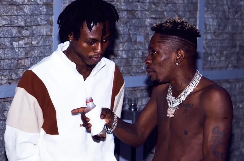 Ghanaian music artists Kweku Flick and Shatta Wale are captured in a candid conversation indoors. Shatta Wale, seen shirtless with his signature heavy silver chains, gestures toward Kweku Flick who is wearing a white and brown tracksuit jacket while holding a bottle wrapped in a white napkin.